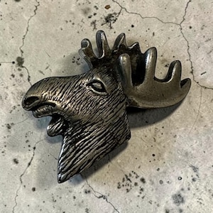 May include: A metallic silver-toned pin shaped like the head of a moose, featuring detailed textured lines that mimic fur and a prominent set of antlers. The object has a polished finish with darker recessed areas that highlight the facial features.