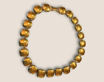 May include: A gold-toned necklace featuring square, textured links. The necklace is composed of multiple square-shaped links connected by smaller, gold-colored connectors. The clasp is also gold-toned.