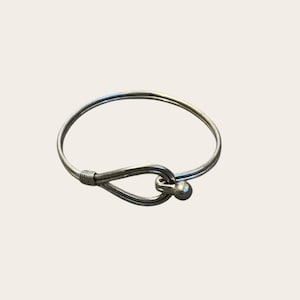 May include: A silver-colored bracelet with a hook and eye closure. The bracelet is a simple, circular design with a small ball at the end of the hook. The hook is wrapped with wire.