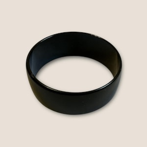 May include: A solid black bangle bracelet. The bracelet is a thick, rounded band with a smooth, glossy finish. The jewelry is a simple, yet elegant accessory.