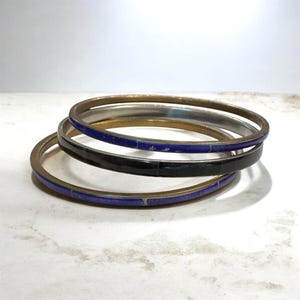 May include: A stack of four metal bangles. One is silver, one is black, and two are gold-toned with blue enamel inlays. The bangles are stacked on a white surface, creating a layered effect. The jewelry is simple and elegant.