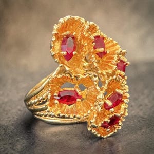May include: A gold-colored ring featuring a cluster of red gemstones. The ring has an ornate, textured design with multiple oval-shaped red stones set within the gold filigree. The band is also textured.