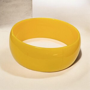 May include: A vibrant yellow bangle bracelet. The bracelet is a solid, smooth band of yellow, with a rounded shape. The bangle is a bold, simple accessory.