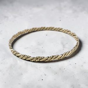 Puede incluir: Una pulsera tipo brazalete circular y delgada, fabricada con un material metálico de tono dorado. La superficie de la pulsera presenta una textura de cuerda trenzada continua y repetitiva que recorre toda la circunferencia del aro.