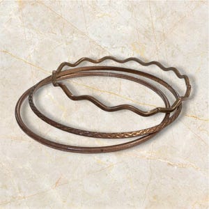 May include: Three bronze bangles. One has a wavy design, another is smooth, and the third features a woven pattern. The bangles are stacked on a light-colored surface.