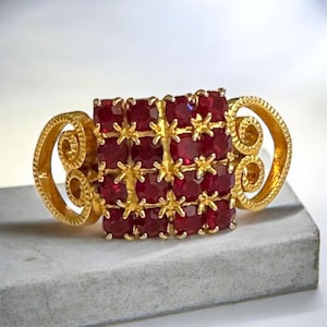 Op de afbeelding: Een goudkleurige ring met een rechthoekige cluster van vierkant geslepen, dieprode edelstenen. De ring heeft sierlijke, wervelende gouden accenten aan de zijkanten en tussen de edelstenen. De ring ligt op een grijze ondergrond.