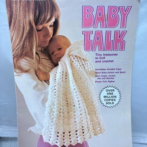 May include: A Leisure Arts craft leaflet titled Baby Talk, featuring a photograph of a person holding an infant wrapped in a white crocheted hooded cape. The cover includes text listing patterns like Snowflake Hooded Cape and Dream Puff Afghan.