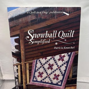 May include: A softcover book titled Snowball Quilt Simplified by Patricia Knoechel, a Quilt in a Day publication. The cover features a log cabin with icicles and a quilt with a white, dark red, and blue star pattern draped over the wooden railing.