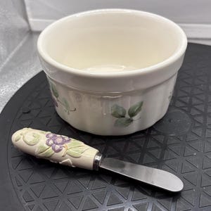 May include: A cream-colored ramekin with a grape and leaf design, paired with a matching butter knife. The knife has a decorative handle with a grape cluster and leaf motif. The ramekin is round and has a slightly fluted edge.