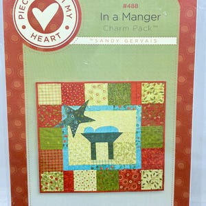 May include: A fabric charm pack titled "In a Manger" by Sandy Gervais. The package features a quilt design with a central manger scene, surrounded by red, green, and patterned fabric squares. The packaging is red and green with the text "Pieces From My Heart".