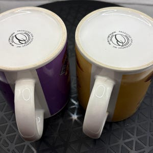 May include: Two ceramic mugs with white handles and rims. One mug is purple, the other is yellow. Both mugs have a white interior and a logo on the bottom. The mugs are likely for beverages.