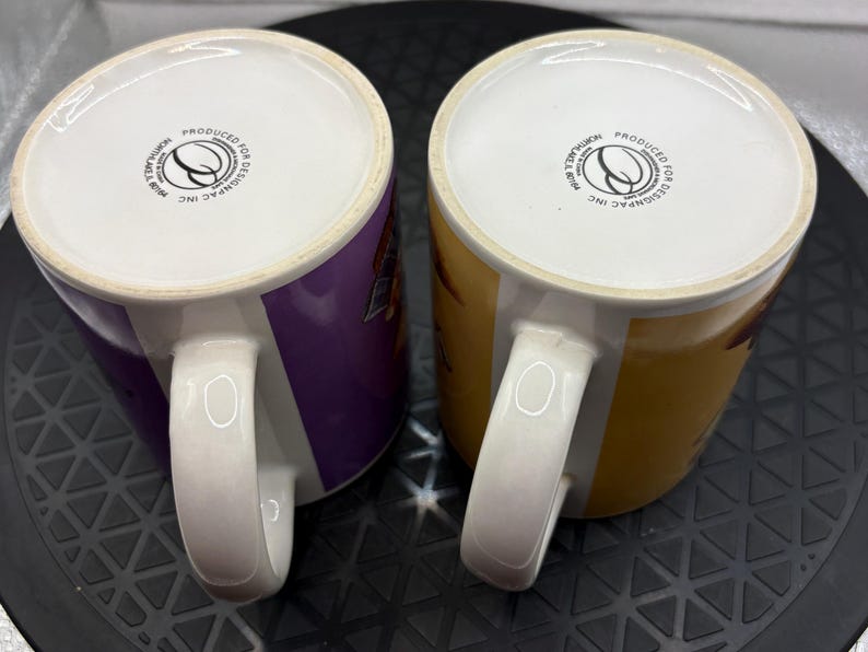 May include: Two ceramic mugs with white handles and rims. One mug is purple, the other is yellow. Both mugs have a white interior and a logo on the bottom. The mugs are likely for beverages.