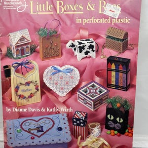 May include: A collection of cross-stitch projects featuring small boxes and decorative items. The image includes a house, a cow, a heart, and a cat-shaped box, all crafted with needlework. The text reads "Little Boxes & B" and "in perforated plastic".