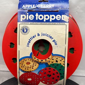 May include: Vintage "pie topper" kit with a blue and white box. The box reads "APPLE/CHERRY" and "prettier & juicier pie." The kit includes red and green pie crust cutters and a pie with a tree-shaped cutout. The box has a red pie-shaped design.