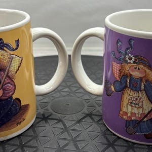 May include: Two ceramic mugs with white handles and rims. One mug is yellow, the other is purple. Each mug features a whimsical illustration of a doll-like figure with wings, holding a tool. The mugs are likely for beverages.