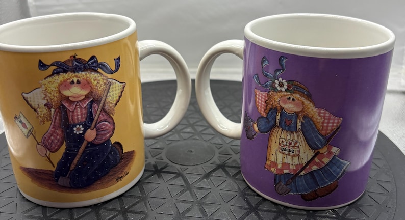 May include: Two ceramic mugs with white handles and rims. One mug is yellow, the other is purple. Each mug features a whimsical illustration of a doll-like figure with wings, holding a tool. The mugs are likely for beverages.