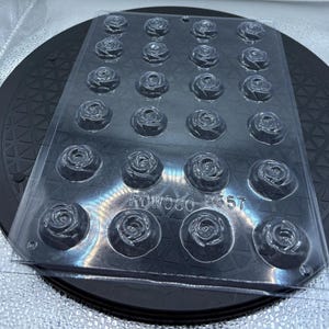 May include: A clear plastic mold with 24 rose-shaped cavities. The mold is rectangular and sits on a black surface. The text "ROWOCO R357" is visible on the mold. Ideal for creating chocolates or candies with a floral design.