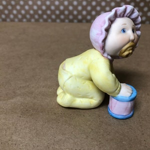 May include: A porcelain figurine of a baby in a yellow onesie and pink bonnet, crawling towards a pink and blue toy drum.