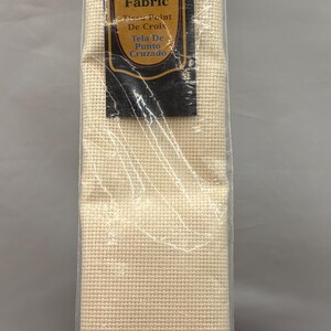 May include: Ivory AIDA cross stitch fabric, 18 count, measuring 15" x 18" (38.1 x 45.7 cm). The fabric is labeled "Gold Quality" and is suitable for cross-stitch projects. The packaging is clear plastic.