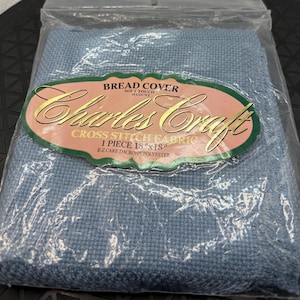 May include: A light blue Charles Craft cross stitch fabric bread cover in a clear plastic package. The label reads BREAD COVER, SOFT TOUCH, 14 COUNT, Charles Craft, CROSS STITCH FABRIC, 1 PIECE 18 inches by 18 inches, E-Z CARE DACRON POLYESTER.