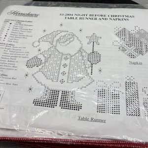 May include: A clear plastic package containing a cross-stitch pattern for a Christmas table runner and napkins. The design features a Santa Claus figure, gifts, and stars. The text "Night Before Christmas" is visible.