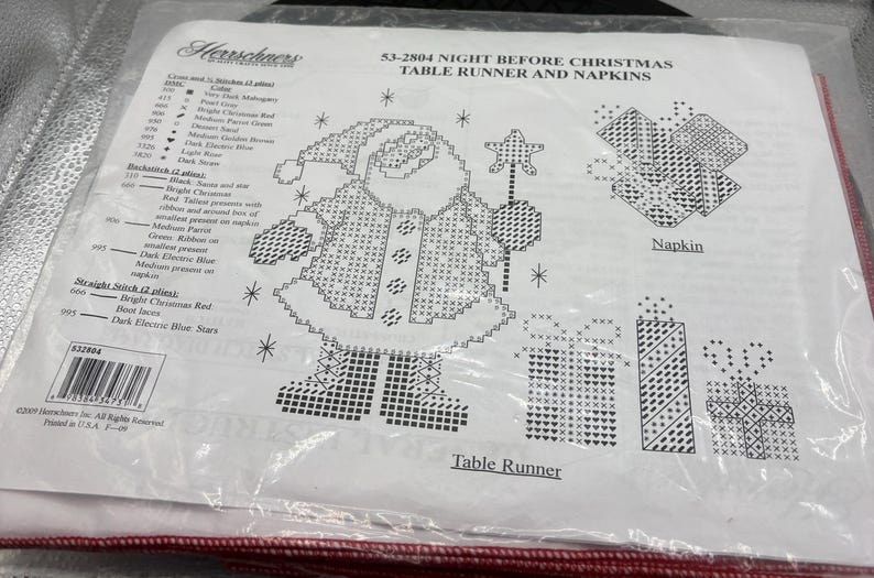May include: A clear plastic package containing a cross-stitch pattern for a Christmas table runner and napkins. The design features a Santa Claus figure, gifts, and stars. The text "Night Before Christmas" is visible.