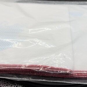 May include: A clear plastic bag containing white fabric and a red cord along the bottom edge. The fabric has blue and white patterned accents. The bag is sealed and appears to be new.