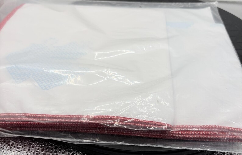 May include: A clear plastic bag containing white fabric and a red cord along the bottom edge. The fabric has blue and white patterned accents. The bag is sealed and appears to be new.