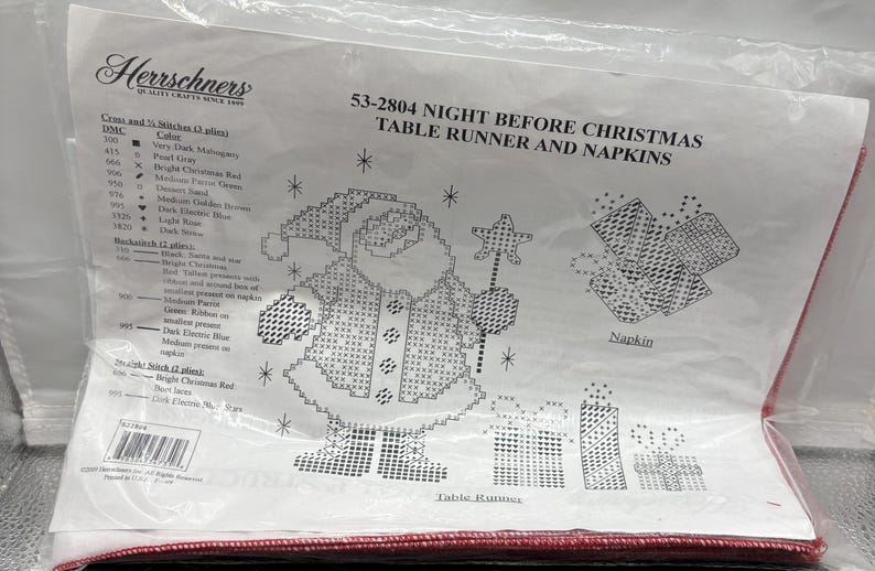 May include: A plastic-wrapped craft kit for a Christmas table runner and napkins. The design features a pixelated Santa Claus holding a star wand, gifts, and snowflakes. The kit includes a color chart and instructions.