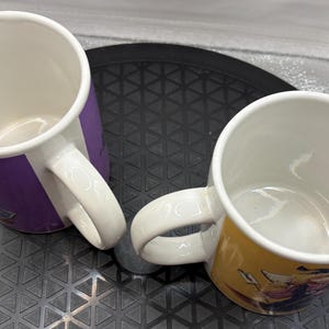 May include: Two ceramic coffee mugs with white interiors and handles. One mug has a purple exterior, the other is yellow. Both mugs have a cartoon graphic design on the exterior. The mugs are on a black surface.