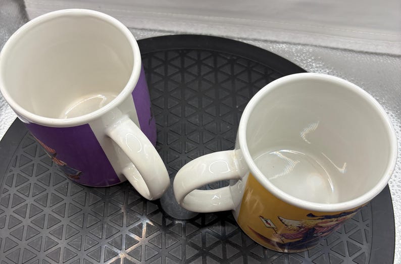 May include: Two ceramic coffee mugs with white interiors and handles. One mug has a purple exterior, the other is yellow. Both mugs have a cartoon graphic design on the exterior. The mugs are on a black surface.