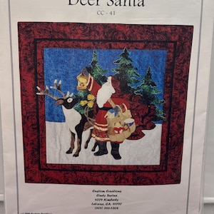 May include: A fabric panel featuring a festive scene of Santa Claus riding a reindeer through a snowy landscape, with evergreen trees in the background. The panel is framed with a red border and includes the text "Deer Santa" and "CC-41".