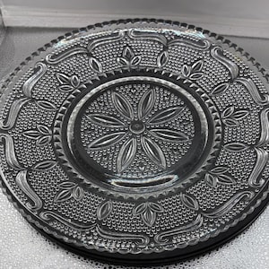 May include: A clear pressed glass plate featuring a central starburst pattern surrounded by a stippled texture. The rim and border are decorated with embossed leaf motifs and curved lines, finished with a scalloped edge design.