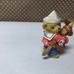 May include: A small, brown teddy bear figurine wearing a sailor hat and red and blue clothing. The bear is holding a brown rocking horse with a red bridle.