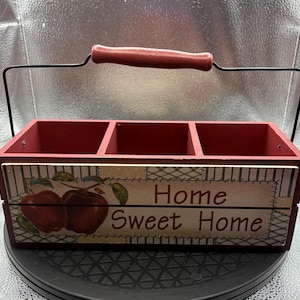 May include: Red wooden caddy with three compartments and a black wire handle with a red wooden grip. The front features a painted design of apples and the words "Home Sweet Home."
