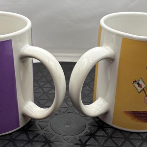 May include: Two white ceramic mugs with curved handles. One mug has a purple background with a cartoon figure, the other has a yellow background with a similar figure. The figures are dressed in whimsical outfits.
