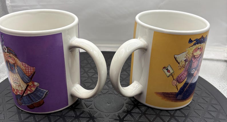 May include: Two white ceramic mugs with curved handles. One mug has a purple background with a cartoon figure, the other has a yellow background with a similar figure. The figures are dressed in whimsical outfits.