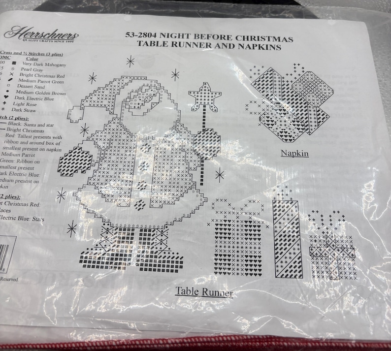 May include: A cross-stitch kit for a Christmas table runner and napkins. The design features Santa Claus, presents, and stars. The kit includes instructions and materials for creating a festive holiday table setting.