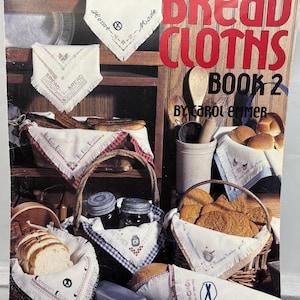 May include: A book cover titled "Bread Cloths Book 2" with various bread cloths displayed. The cloths are white with embroidered designs and text, some in baskets with bread. The cover has a wooden background with text and a price.