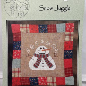 May include: A cross-stitch pattern titled "Snow Juggle" featuring a snowman with a carrot nose and a red and white scarf. The design is framed by a patchwork border of red, blue, and plaid fabrics. The chart number is KTN033.