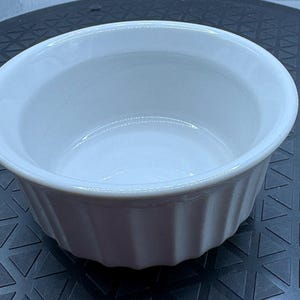 May include: A white ceramic ramekin with a fluted exterior. The small, round dish has a smooth interior and is designed for baking and serving individual portions of food.