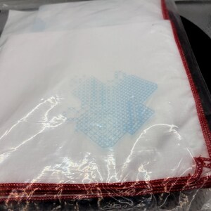 May include: A stack of white handkerchiefs with a blue design and red stitched edges, wrapped in clear plastic. The handkerchiefs appear to be folded and stacked neatly, ready for use.
