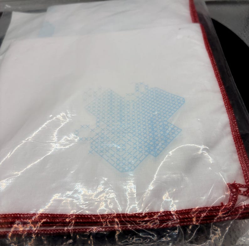 May include: A stack of white handkerchiefs with a blue design and red stitched edges, wrapped in clear plastic. The handkerchiefs appear to be folded and stacked neatly, ready for use.