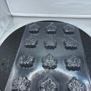 May include: Clear plastic mold with 12 maple leaf-shaped cavities. The mold is designed for creating decorative items, possibly chocolates or soaps. The cavities are detailed with leaf vein patterns.