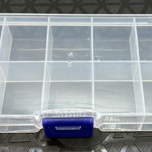 May include: A clear plastic organizer with multiple compartments. The rectangular box has a blue clasp and a small loop on the side. The interior is divided into several rectangular sections, ideal for storing small items.