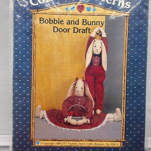 May include: A package for a door draft pattern featuring two bunny figures. The bunnies are dressed in red outfits with white accents. The text on the package reads "Country Patterns" and "Bobbie and Bunny Door Draft."