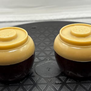 May include: Two vintage salt and pepper shakers. Each shaker has a dark brown base and a tan top with a small, circular design. The shakers are round and have a classic, retro aesthetic.