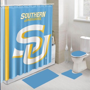 May include: A blue shower curtain with a white and yellow Southern Jaguars logo. The shower curtain is hanging in a bathroom with a white toilet and a blue toilet seat.