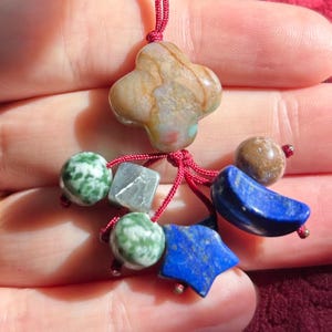 May include: A handcrafted pendant featuring a four-leaf clover-shaped stone with brown and beige hues, accented by a variety of beads. The beads include green speckled spheres, a gray square, a blue crescent, a blue star, and a brown sphere, all strung on a red cord.