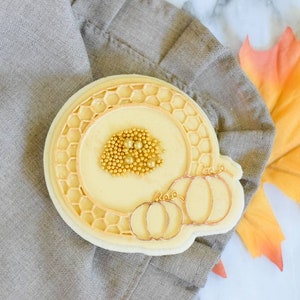 Op de afbeelding: Een witte koek versierd met een honingraatontwerp en gouden sprinkles. De koek heeft ook een ontwerp van drie pompoenen met stelen.